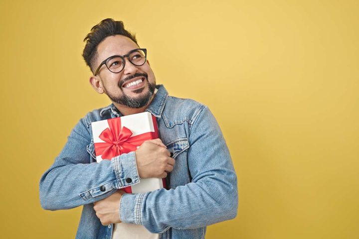 Young man in denim jacket smiling and hugging a gift with a red ribbon on it.