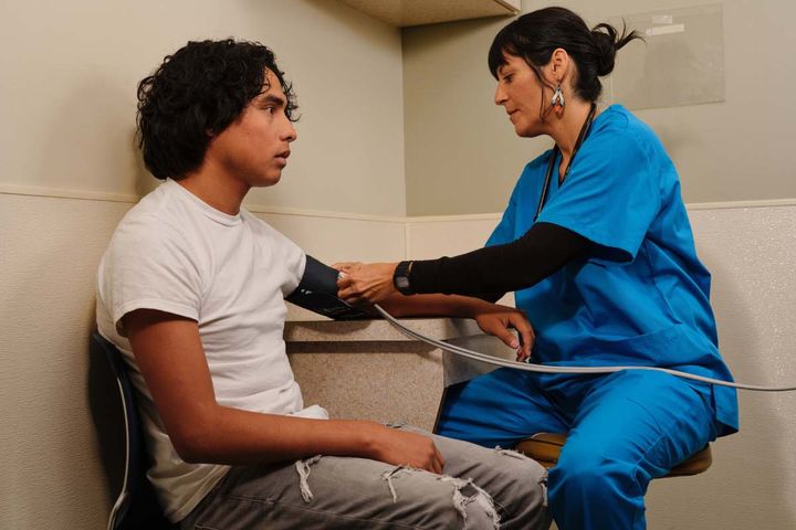Female ER tech in blue scrubs taking blood pressure of young man in a white t-shirt in a corner of the emergency department.