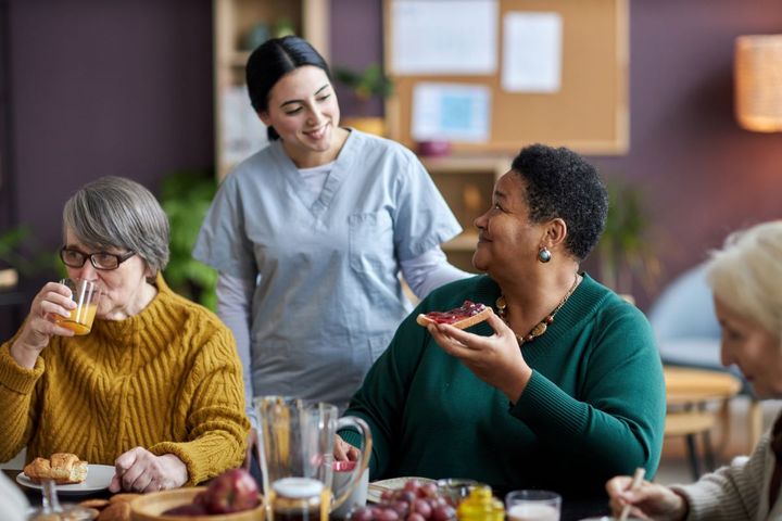 A CNA checks on residents of an LTC facility who are eating breakfast.