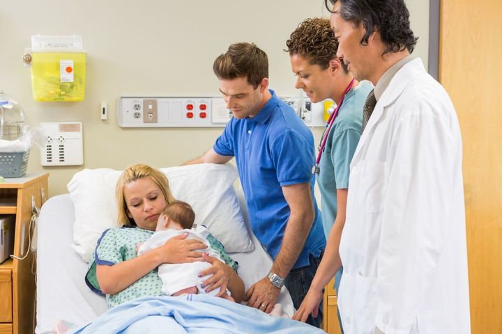 A new mom holds her baby as her partner, the doctor, and postpartum nurse look on.