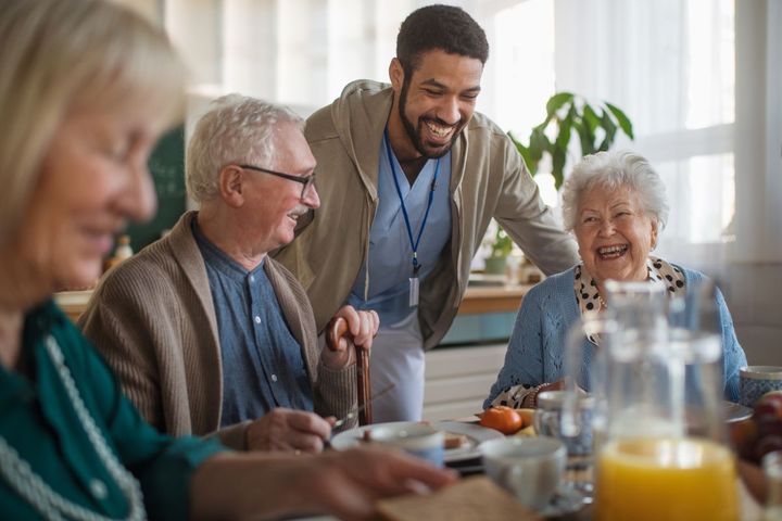 A nurse stands beside a group of nursing home residents, who are laughing as they share a meal together.