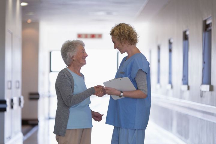 An unlicensed assistive personnel speaks with a patient in a nursing home hallway.