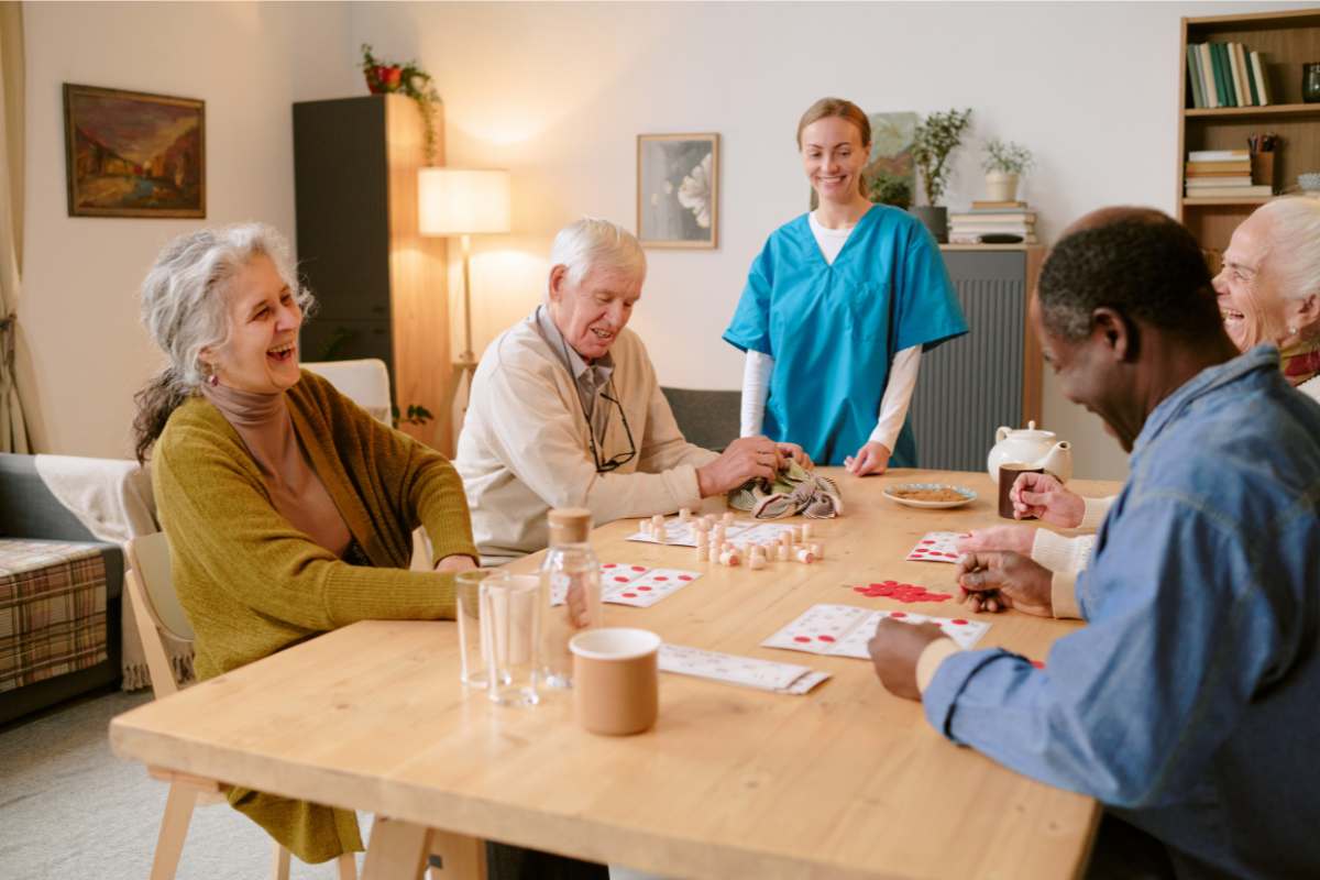 Nurse in scrubs watching a table of happy seniors at an assisted living center play cards.