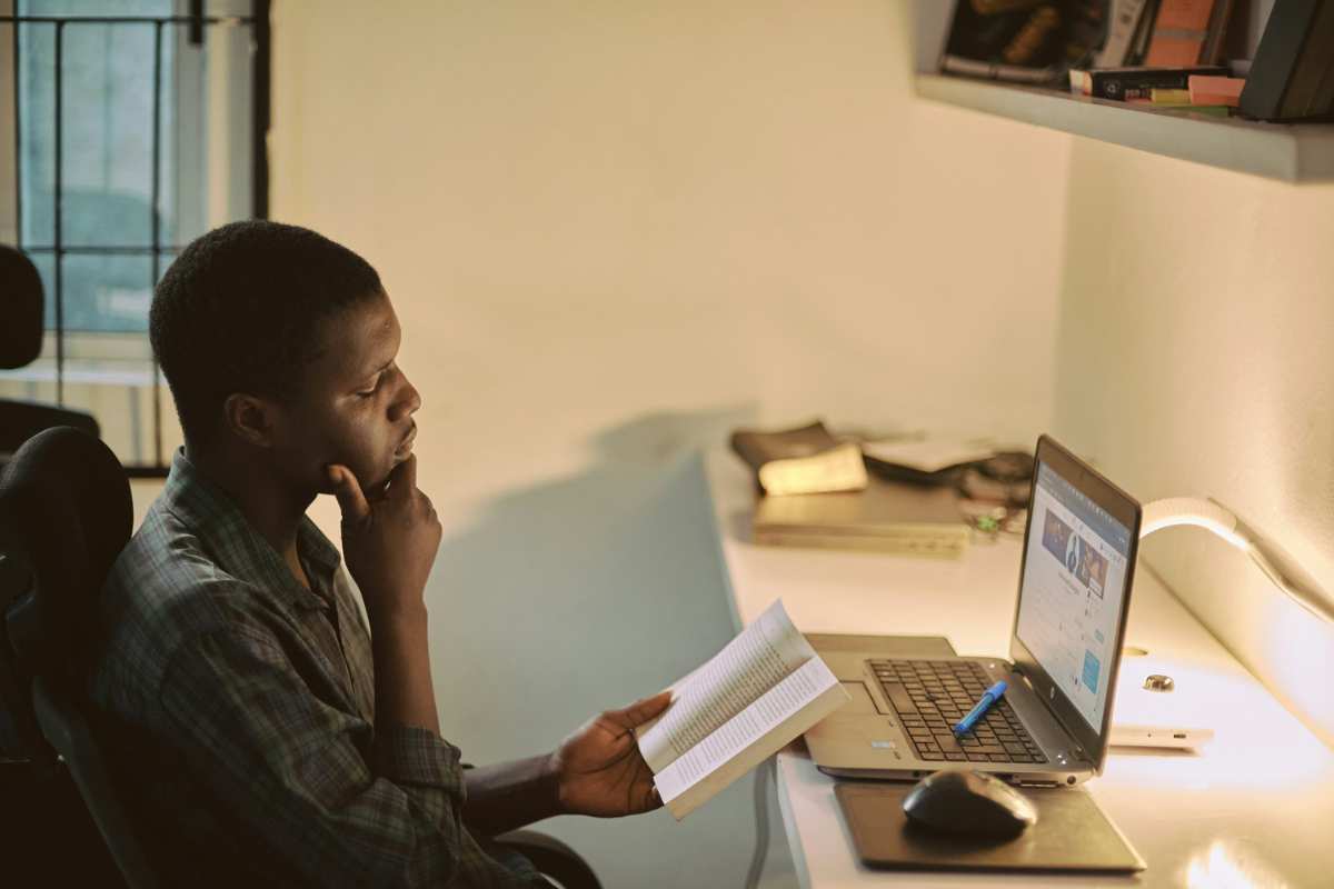 Young Black man reading a book and taking an NCLEX preparation course on laptop.