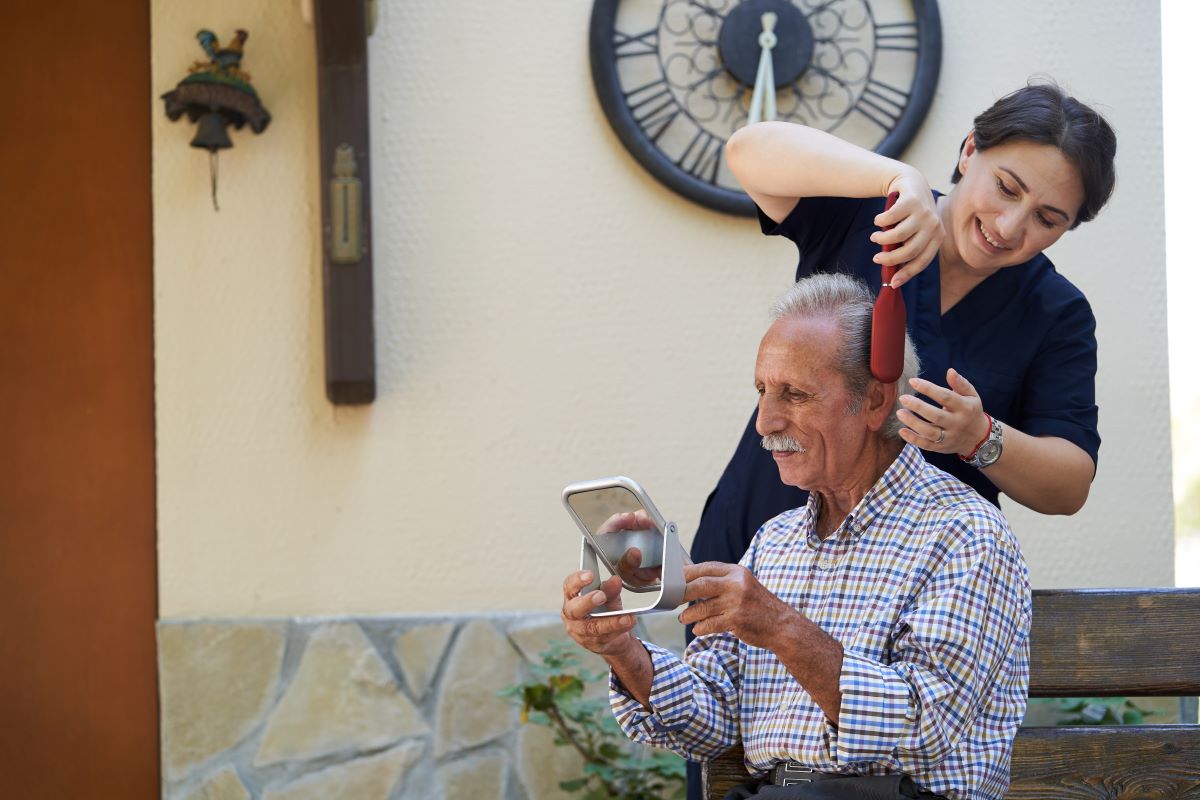 A caregiver at a nursing home brushes a resident's hair as he looks on with a handheld mirror.