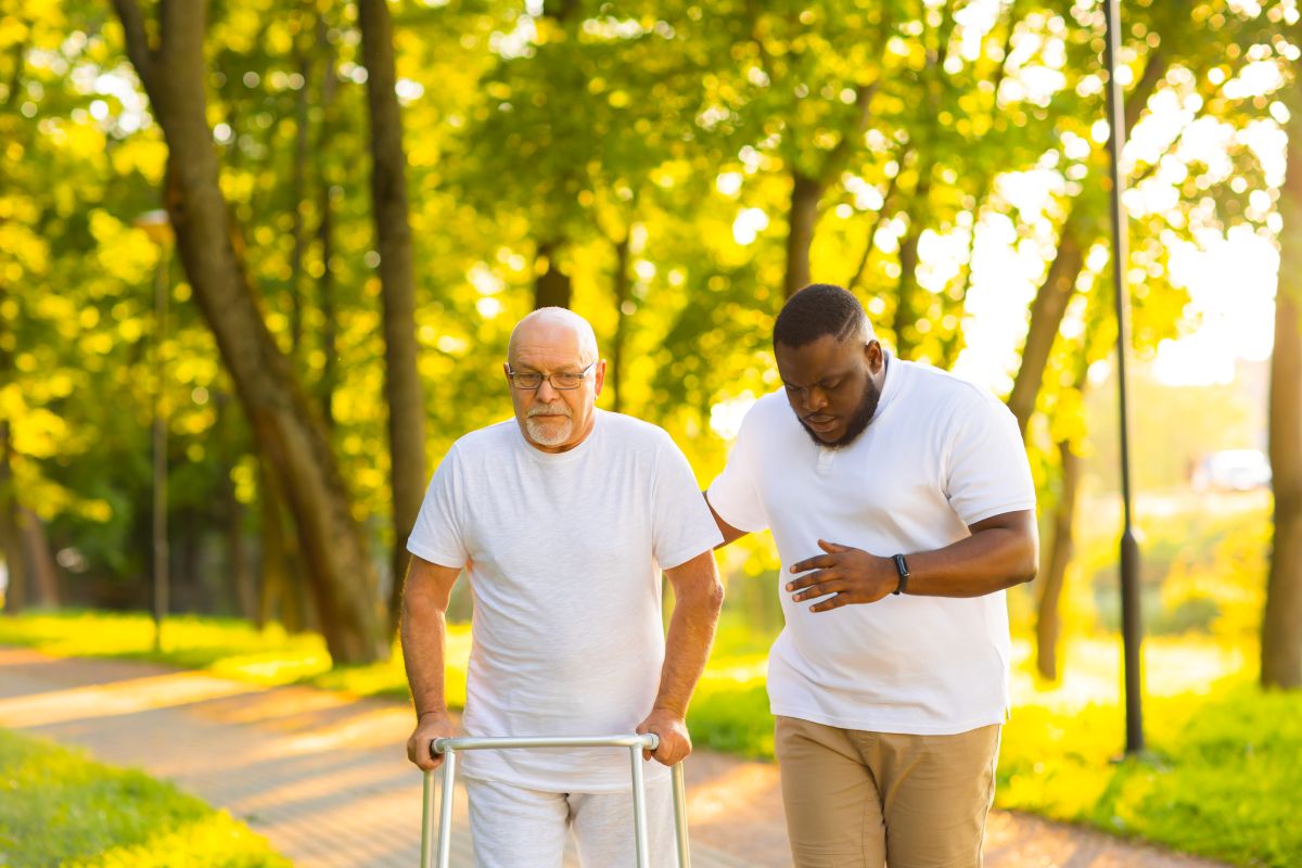A caregiver assists an elderly man as he goes for a walk outside.