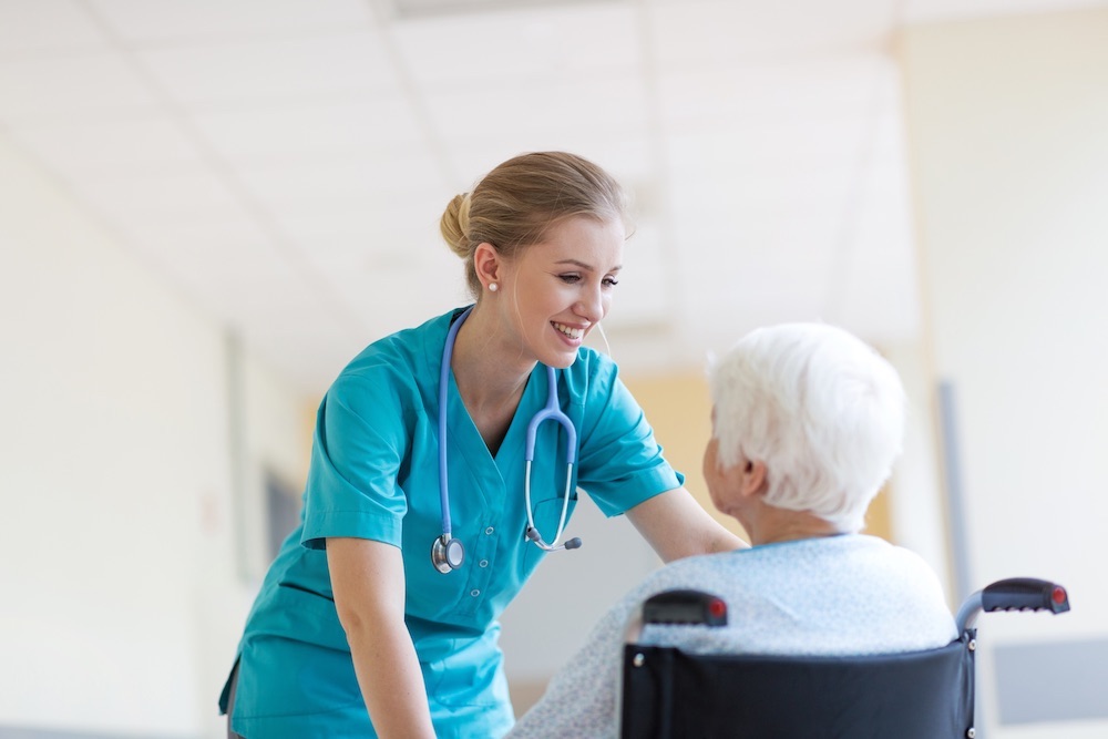 Young nurse with teal scrubs smiling and assisting a senior in a wheelchair.