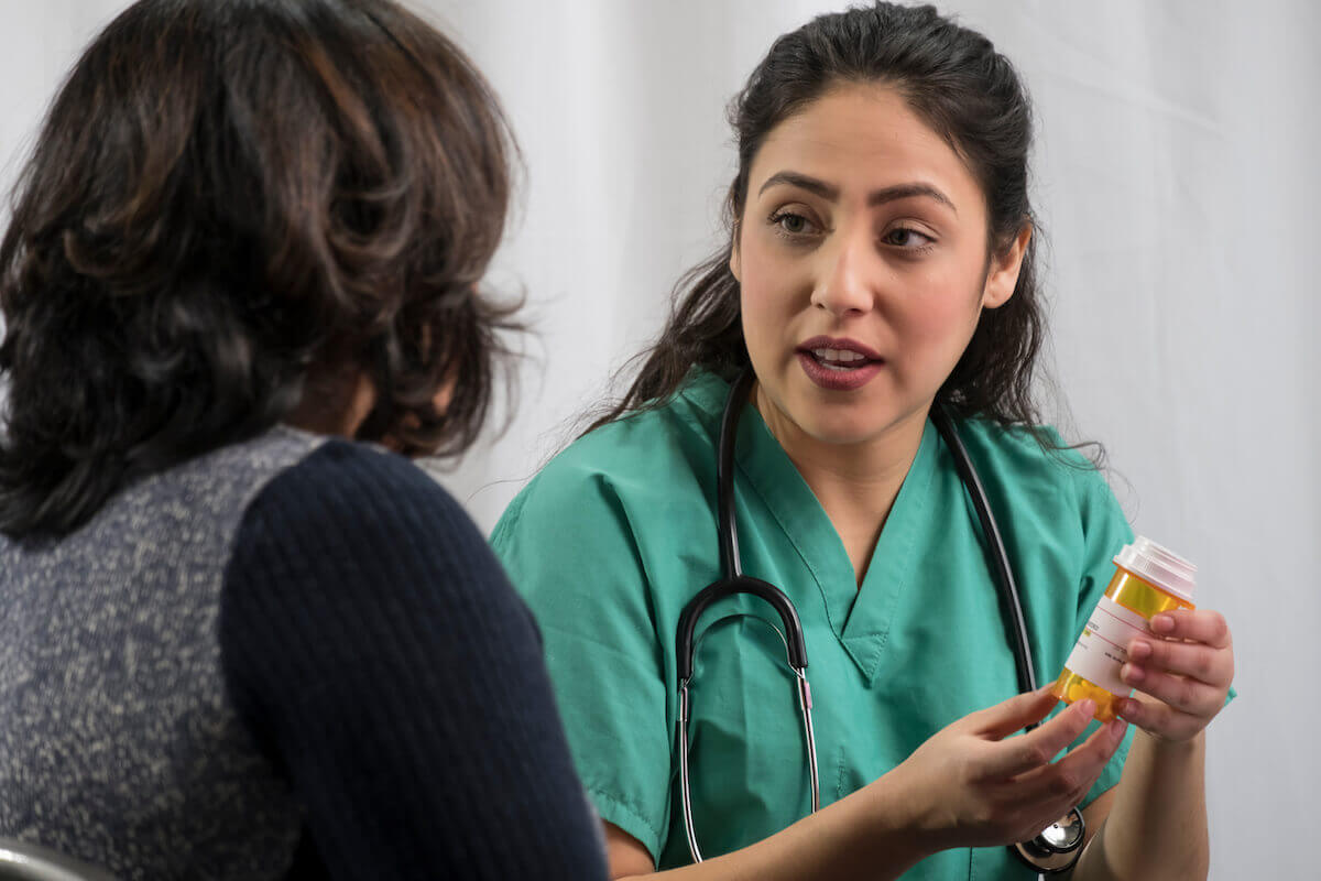 Nurse with long hair in green scrubs holding a medication bottle and talking to a patient.