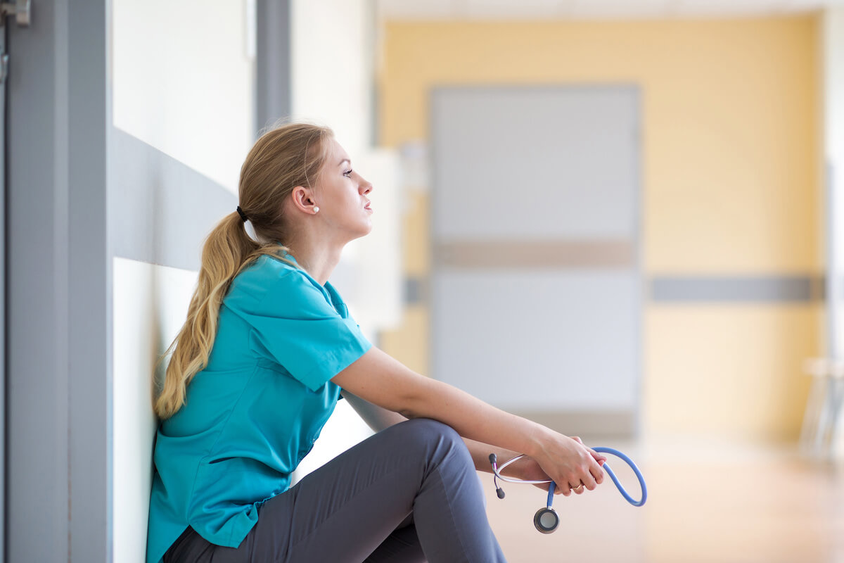 Nurse with long blonde hair sitting in hallway looking forlorn.