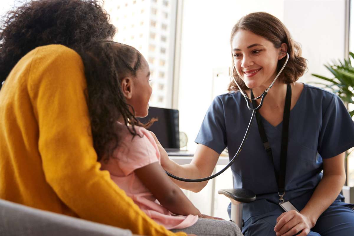 Young female nurse listening to heart of pediatric patient sitting on adult's lap.